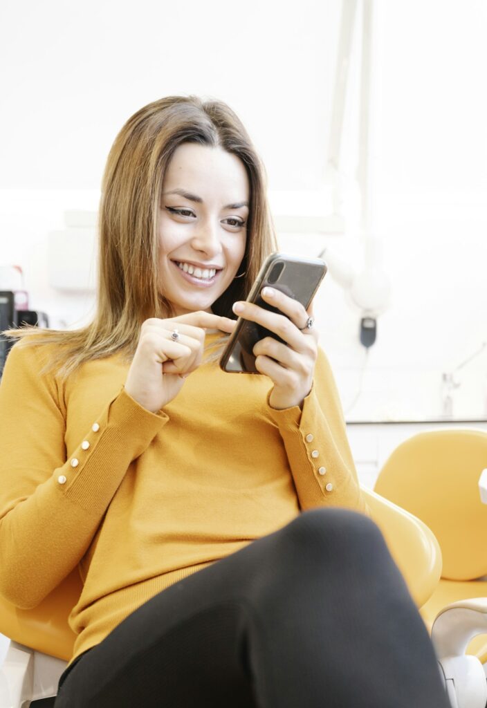 Cheerful woman wearing a yellow sweater using a phone in the dental clinic, displaying patient satisfaction